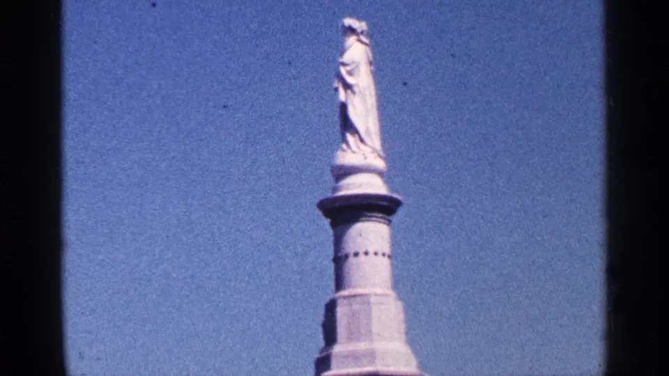 1964 Pennsylvania home-movie frame showing the Gettysburg monument statue on a column, photographed against blue sky.