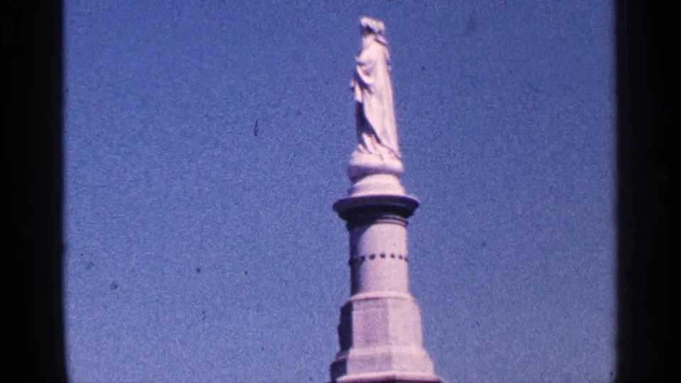 Visitor-shot 8mm view of the monument's upper statue and column, with dark film borders and clear sky visible.