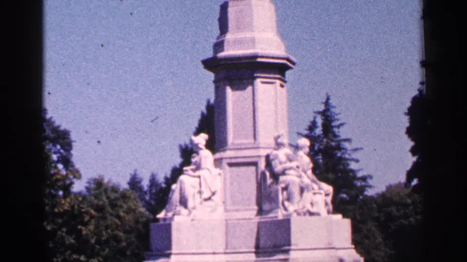 1964 Pennsylvania home-movie frame showing more of the monument, including seated sculptural human figures near the base and trees behind.