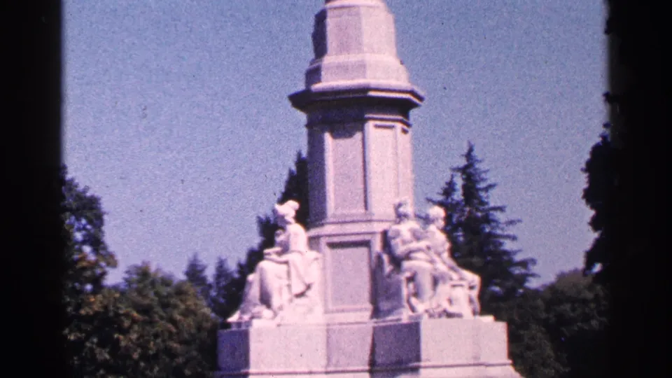 Handheld 8mm frame of a Pennsylvania monument with a tall column, carved seated figures, trees, and bright blue sky.