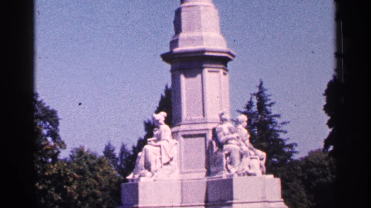 1964 Pennsylvania 8mm frame showing a tall white stone monument with seated and draped sculpted figures under bright sun.