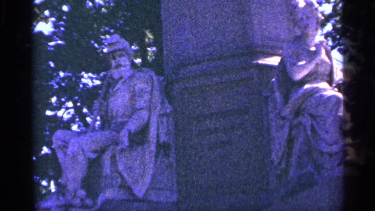 Close view of stone sculpted figures beside the monument base, partly shaded by trees.