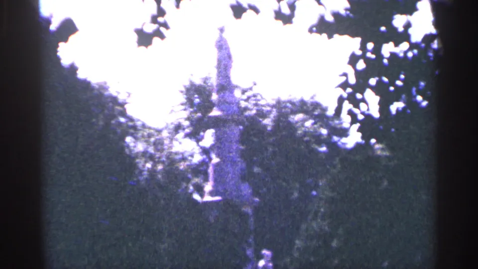 Distant 1964 Pennsylvania view of the monument through foliage, with bright overexposed sky and dark 8mm frame edges.