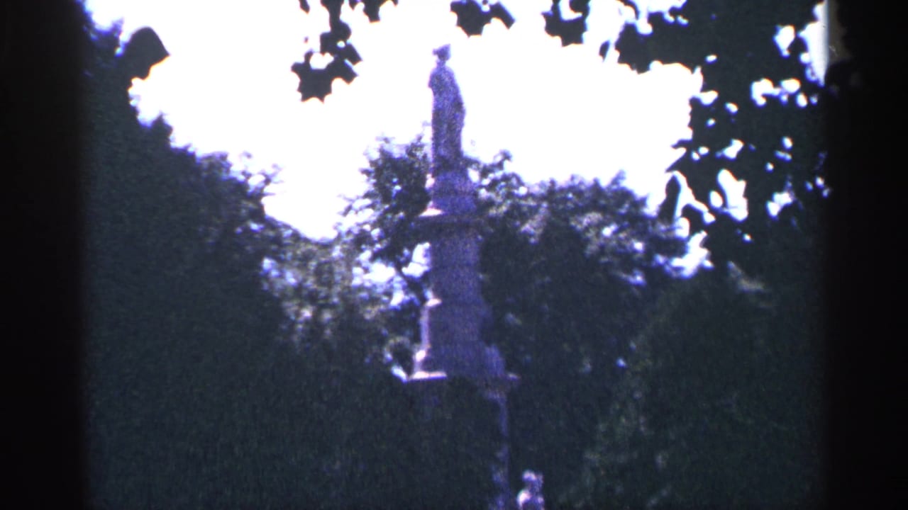 1964 Pennsylvania 8mm frame showing a tall park statue above dense trees, with dark leaves at the top edge and overexposed sky behind it.