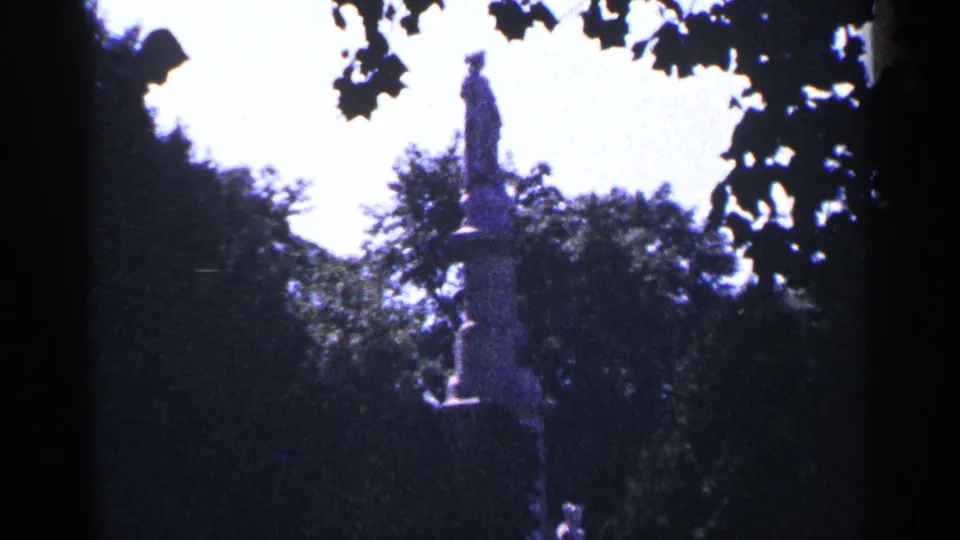1964 Pennsylvania home-movie frame with a distant statue on a pedestal, surrounded by dark green trees and partly framed by overhead leaves.