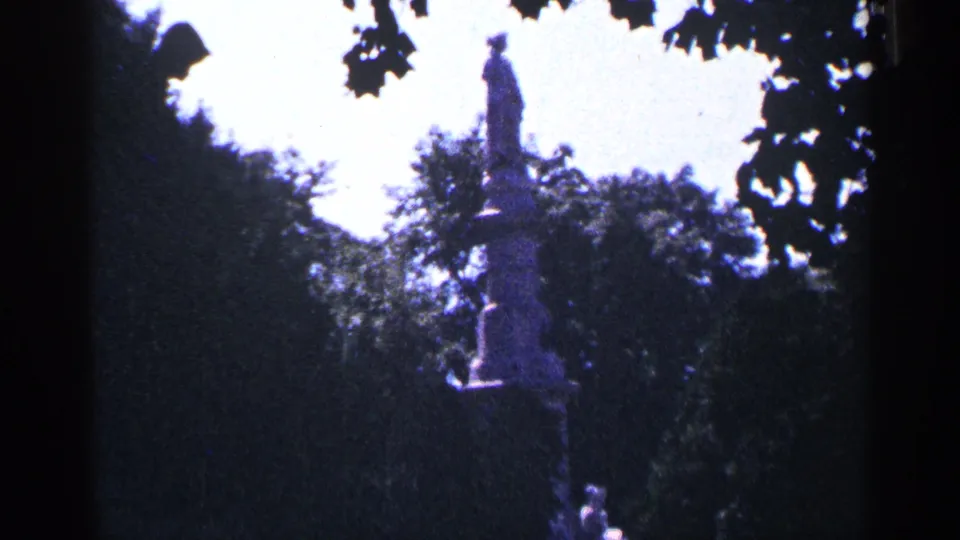 1964 Pennsylvania 8mm view of a tall monument or statue centered beyond park trees, with leafy branches silhouetted against the bright sky.