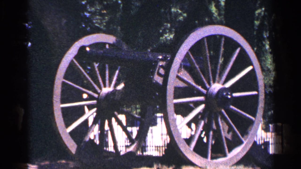Dark historic cannon with large spoked wheels displayed outdoors among trees.
