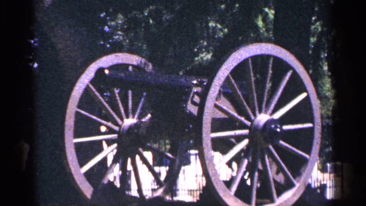 1964 Pennsylvania home-movie frame of a historic wheeled cannon outdoors, with large spokes and shaded trees behind it.