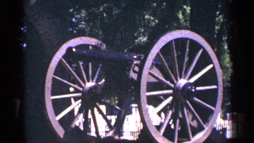 1964 Pennsylvania home-movie frame of a historic wheeled cannon outdoors, with large spokes and shaded trees behind it.