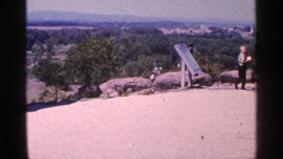 A wider view across the Little Round Top overlook shows rocks, a standing person, a wooden fixture, trees, and distant terrain.