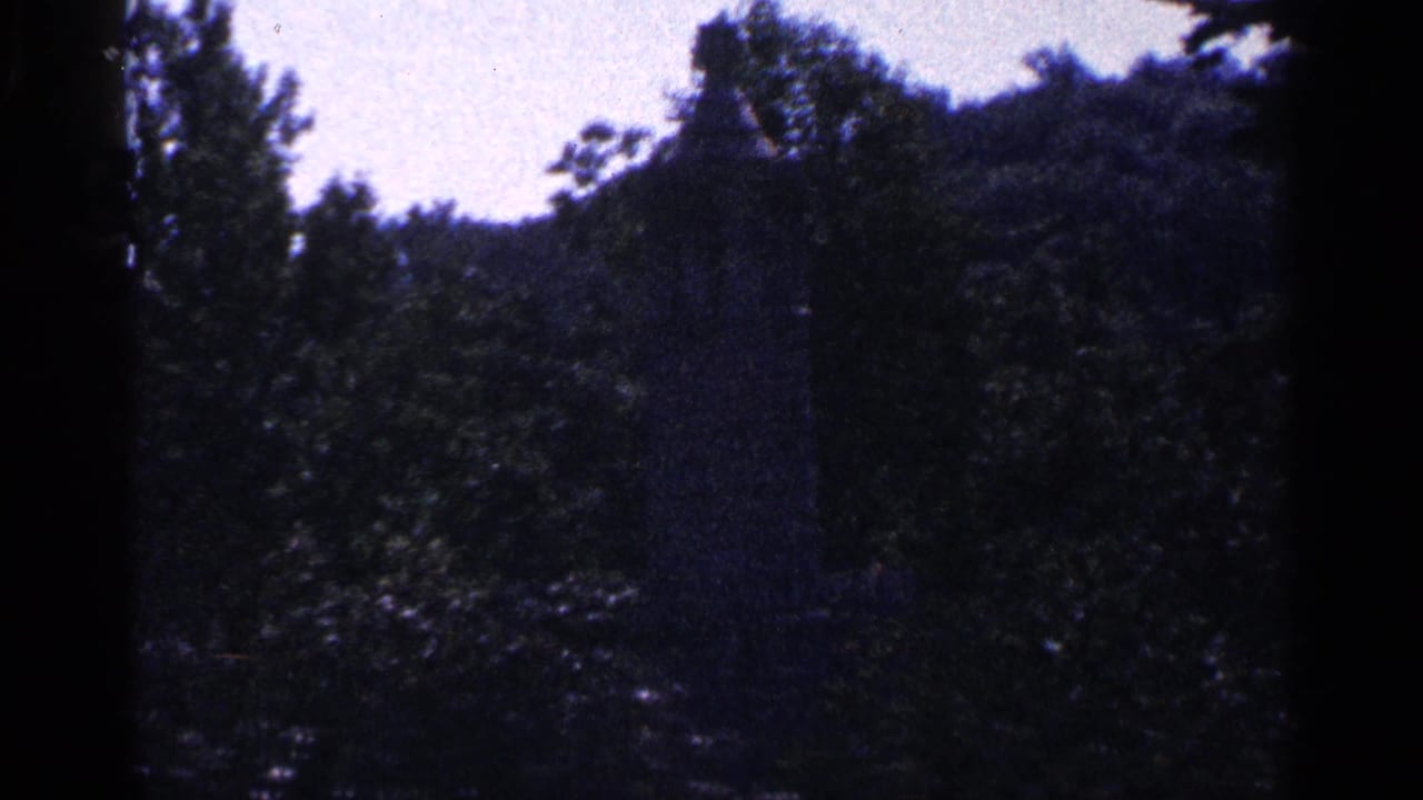 1964 Pennsylvania 8mm frame with a distant grey stone statue or monument partly hidden in dense trees under bright sky.