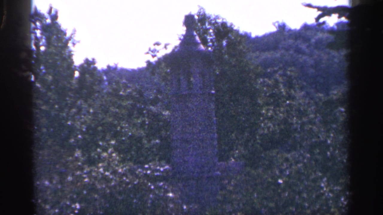 Handheld 1964 Pennsylvania home-movie view of a tall grey statue or monument rising among green trees and hillside foliage.