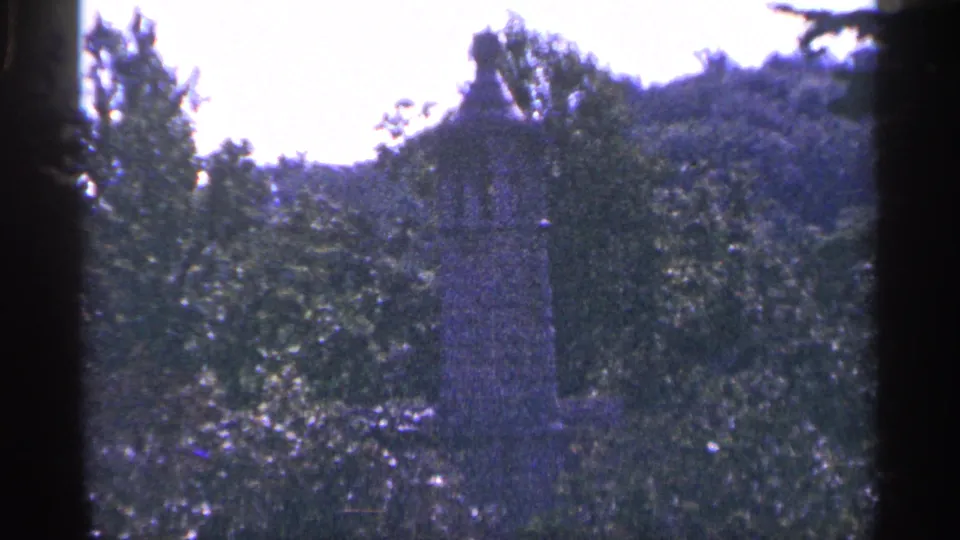 1964 Pennsylvania 8mm shot of a grey statue or monument amid dense foliage, with uneven ground and a wooded hillside behind it.