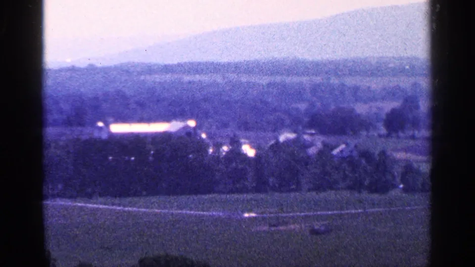1964 Pennsylvania home-movie frame showing open fields, a distant settlement edge, and a blue-purple mountain backdrop.