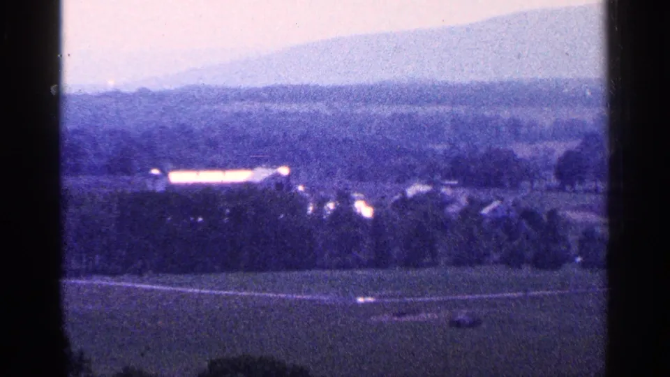 1964 Pennsylvania 8mm frame of farmland, small distant structures, and a soft mountain silhouette under cool film color.
