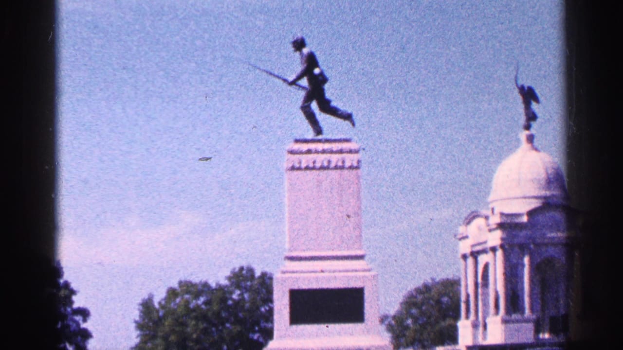Soldier statue on a tall monument beside a domed memorial structure under blue sky.