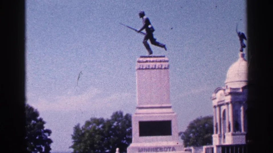 1964 home-movie frame of the First Minnesota Volunteer Regiment Monument at Gettysburg, with the pedestal and nearby domed memorial in daylight.