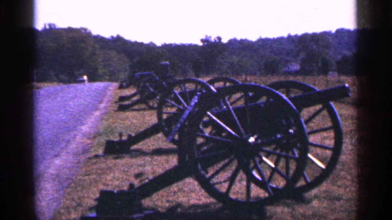 1964 Pennsylvania home-movie frame showing field cannons lined on grass beside a paved road under a bright sky.