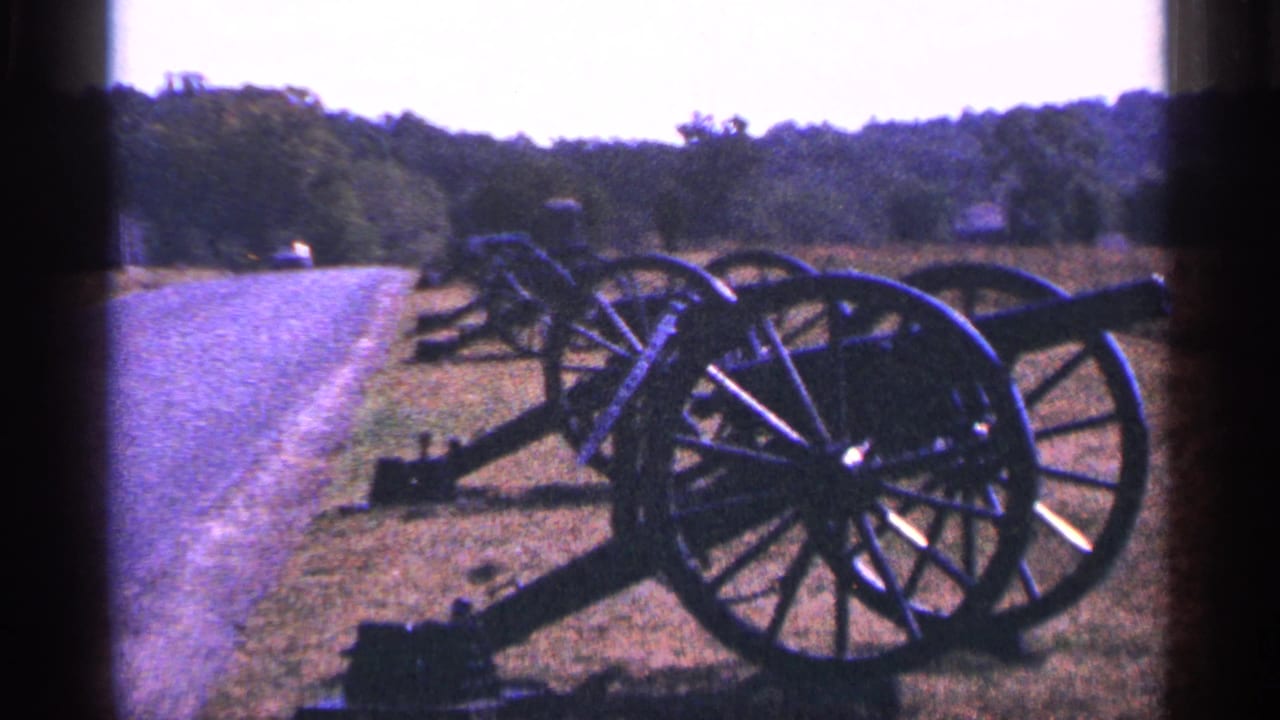 Visitor-shot 8mm frame of historic cannons on grass beside a paved road in Pennsylvania, with trees beyond.