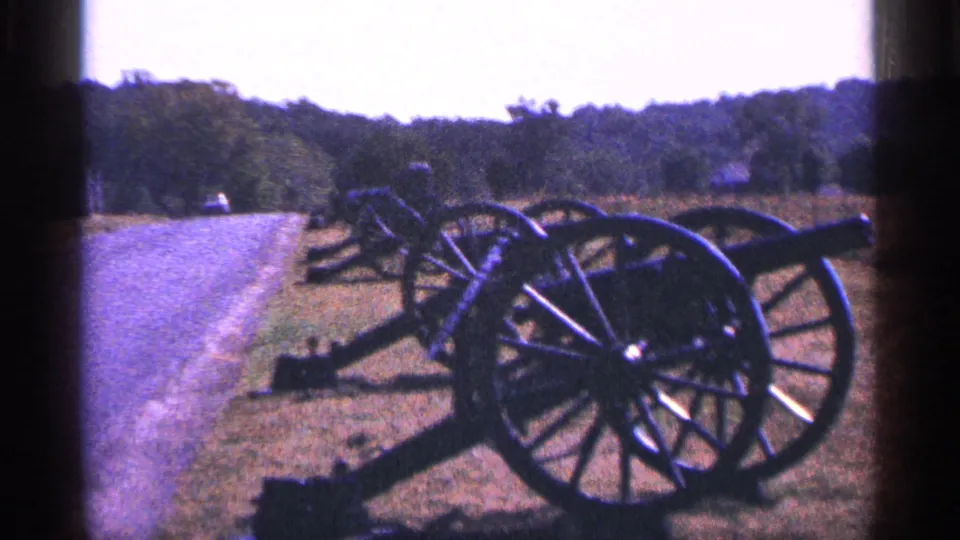 1964 8mm frame showing a closer roadside view of battlefield cannons with grass, road, and trees behind.