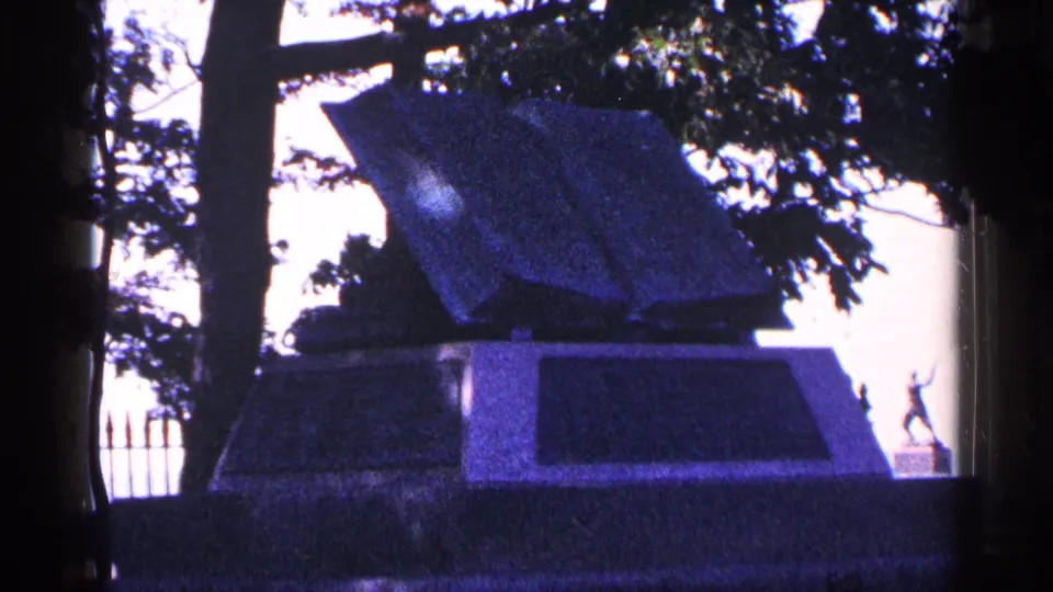 1964 Pennsylvania home-movie frame of a stone memorial or marker shaded by trees, with a small statue visible at right.