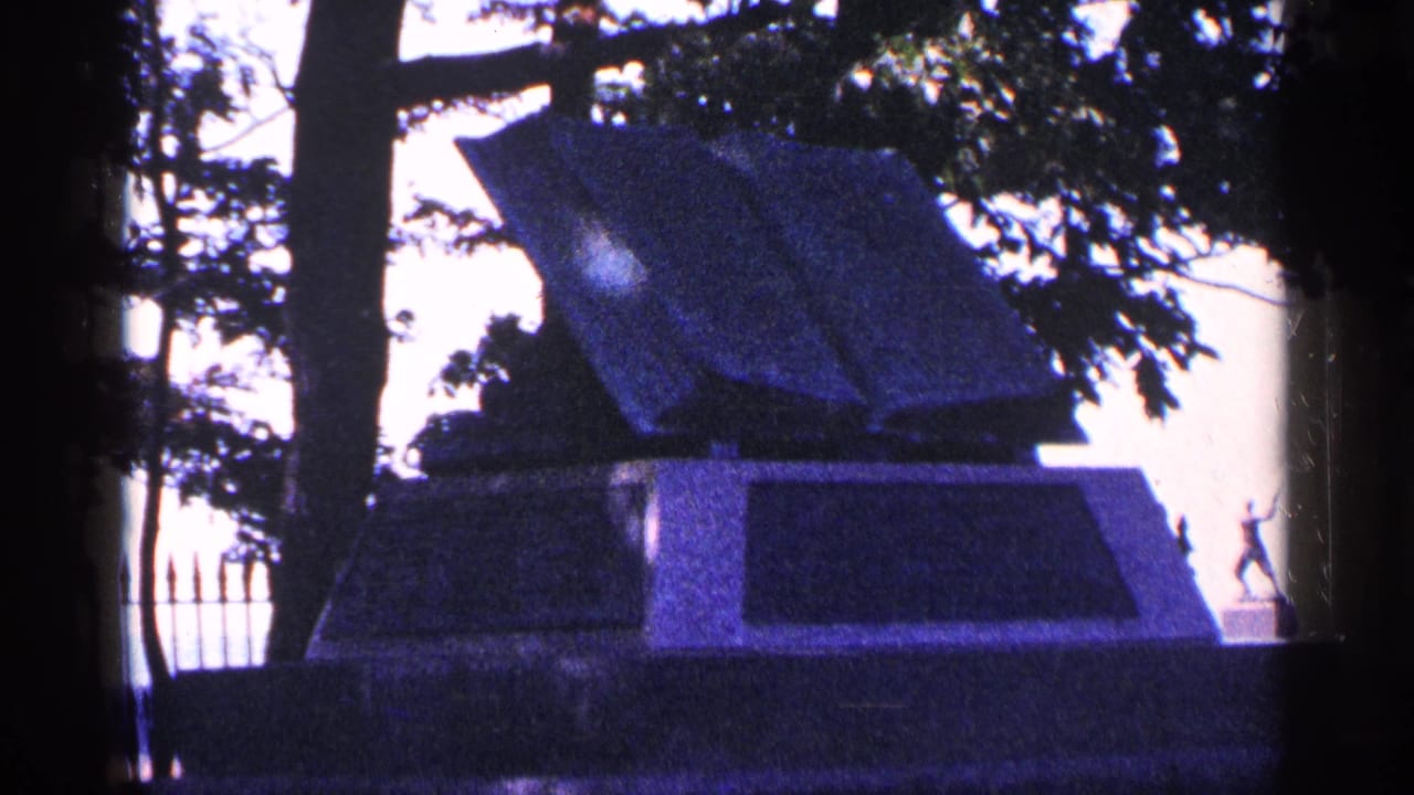 Open book-shaped stone monument framed by trees and a fence.