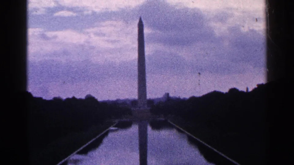 1964 home-movie frame showing the Washington Monument in the distance with its reflection in the Reflecting Pool beneath a cloudy sky.