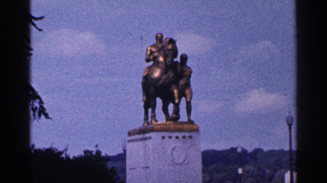 1964 Washington DC home-movie frame showing a distant bronze horse-and-rider monument on a pale pedestal, framed by trees and sky.