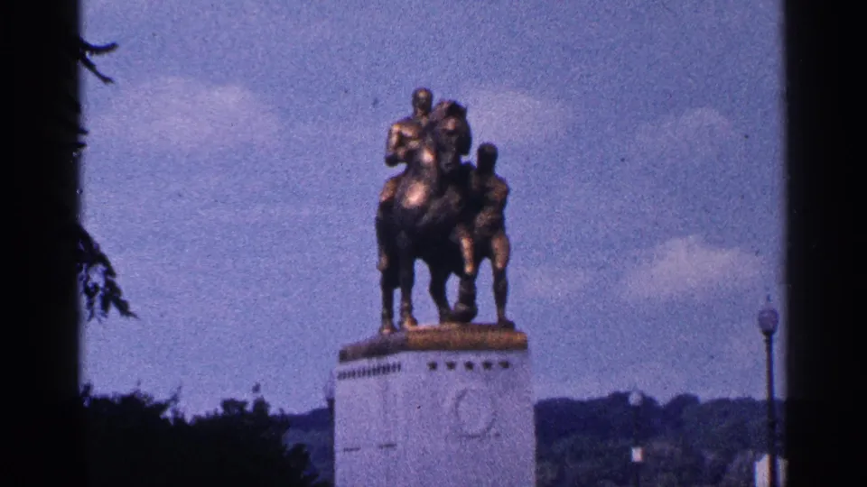 1964 Washington DC home-movie frame showing a distant bronze horse-and-rider monument on a pale pedestal, framed by trees and sky.
