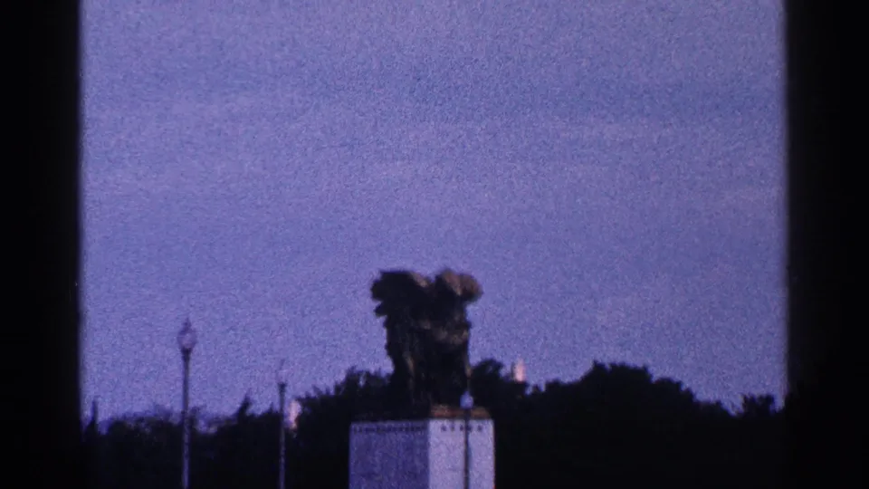 Final distant Washington DC home-movie view with the monument reduced in the frame, set above dark trees under a blue sky.