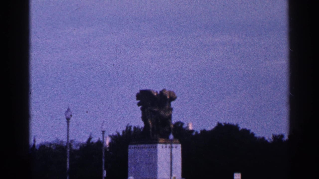 1964 Washington, DC home-movie view of a bronze statue on a white pedestal, framed against trees, lampposts, and open blue sky.