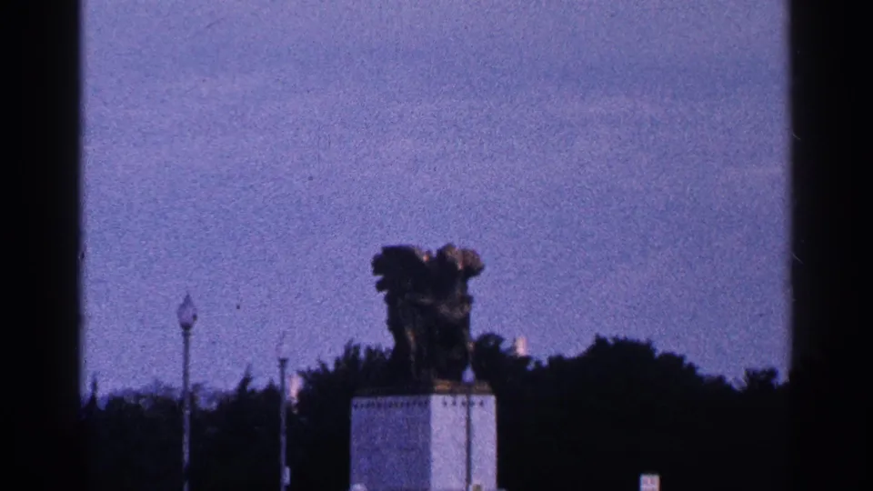 1964 Washington, DC home-movie view of a bronze statue on a white pedestal, framed against trees, lampposts, and open blue sky.