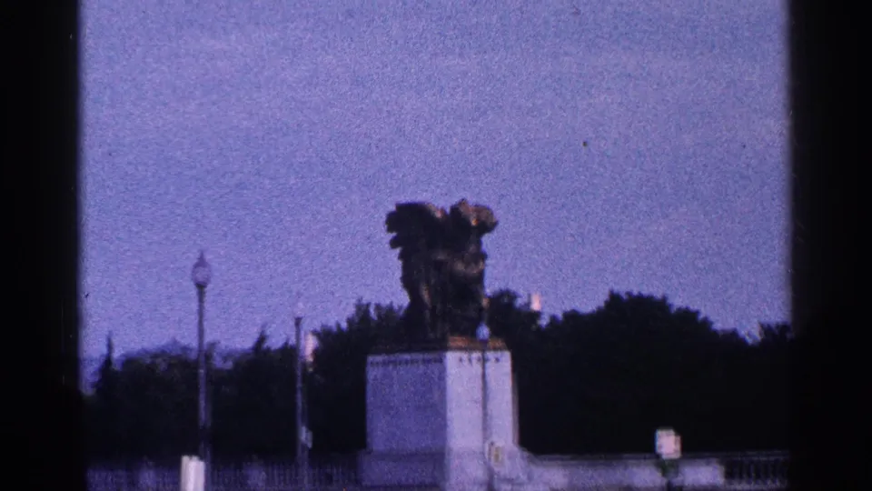 Distant 1964 Washington, DC view of a statue on a white pedestal with dark trees and decorative lampposts in the background.