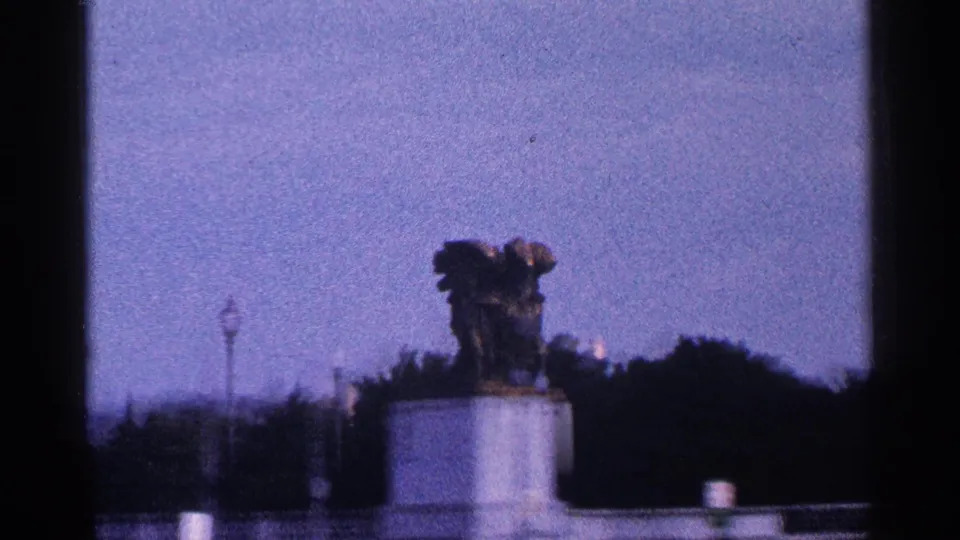 Closer home-movie frame of a bronze statue on a white pedestal in Washington, DC, with trees, lampposts, and open sky visible.