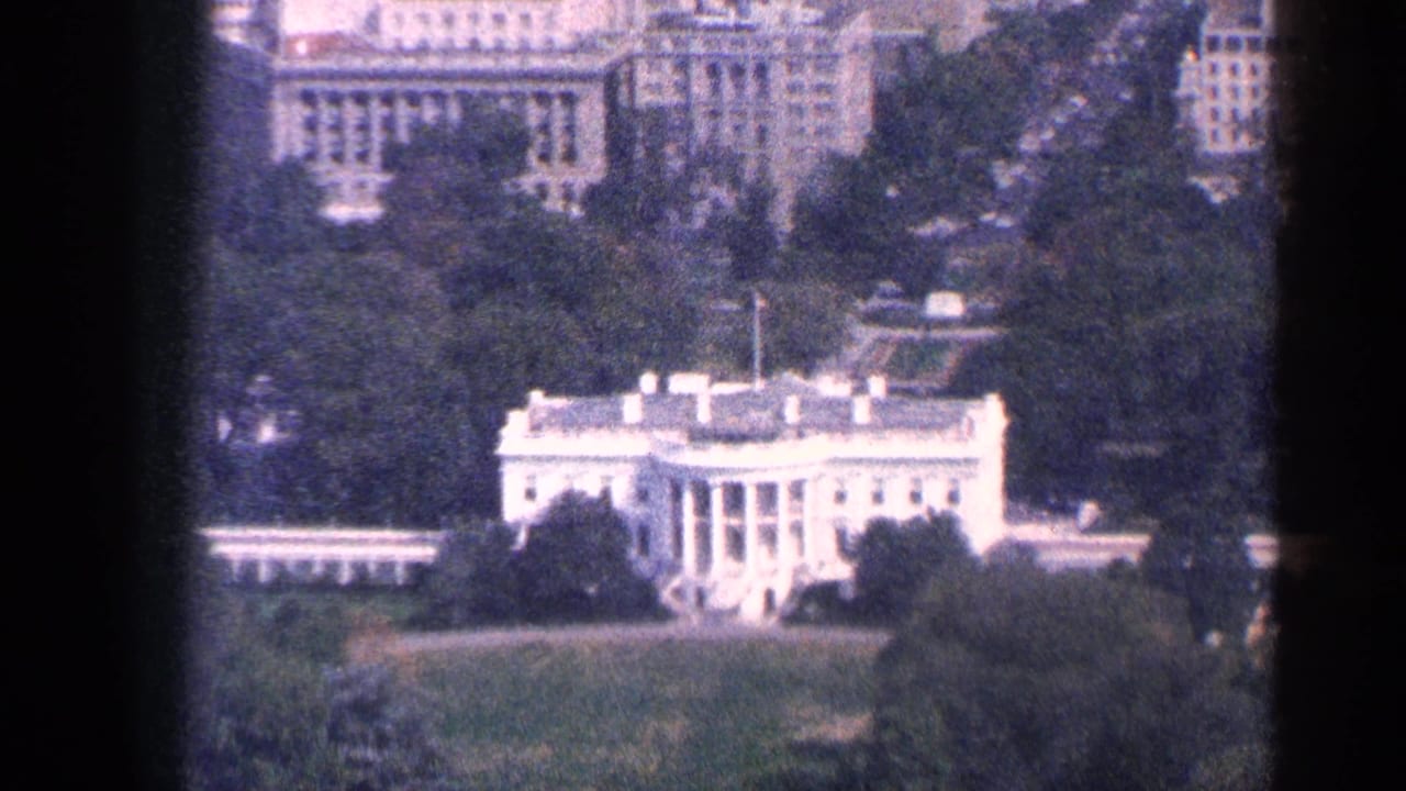 1964 Washington, DC home-movie frame showing the White House from a distant elevated viewpoint, bordered by trees and surrounding buildings.