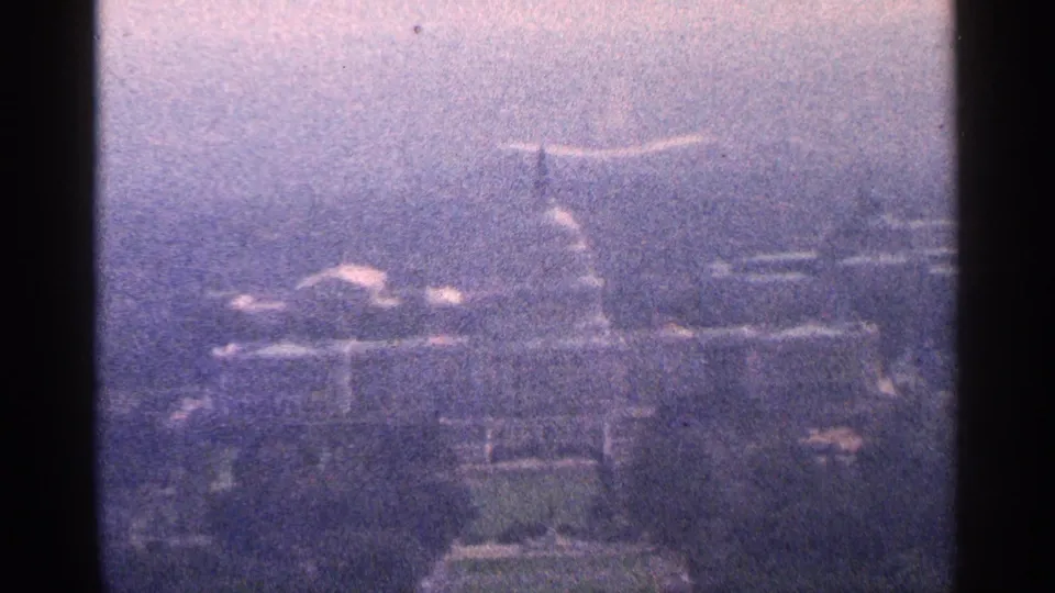 1964 Washington, DC home-movie frame holding on the U.S. Capitol dome and surrounding cityscape, with visible grain, haze, and dark 8mm frame borders.