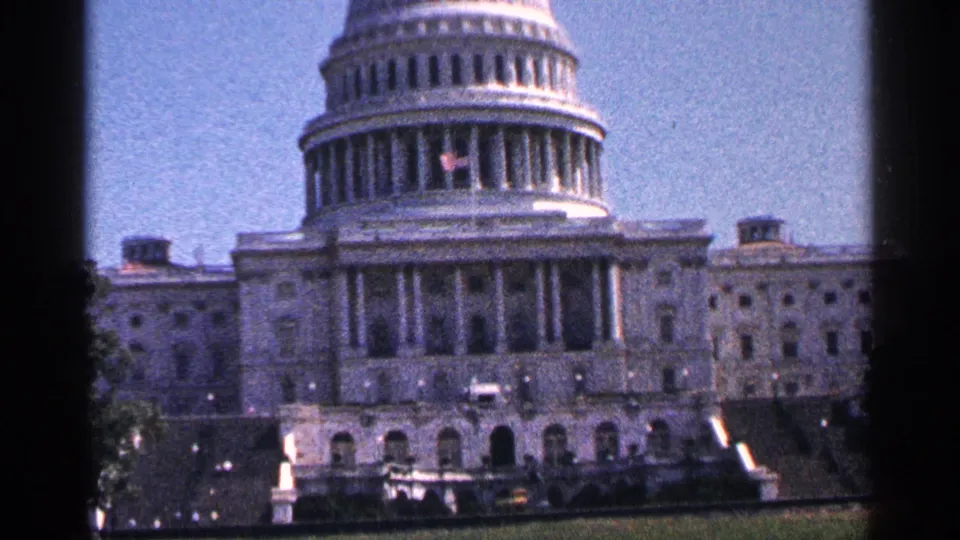 1964 Washington, D.C. home-movie frame of the U.S. Capitol dome and facade, with grain, blue sky, and dark gate edges.
