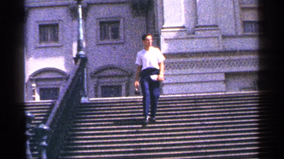 The man moves lower on the Washington DC staircase, framed by railings, arched windows, and bright exterior light.