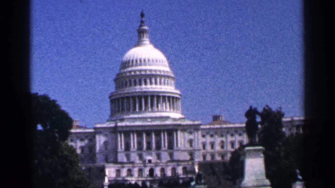 1964 8mm view of the U.S. Capitol in Washington DC, framed by trees, a statue silhouette, blue sky, and dark film edges.