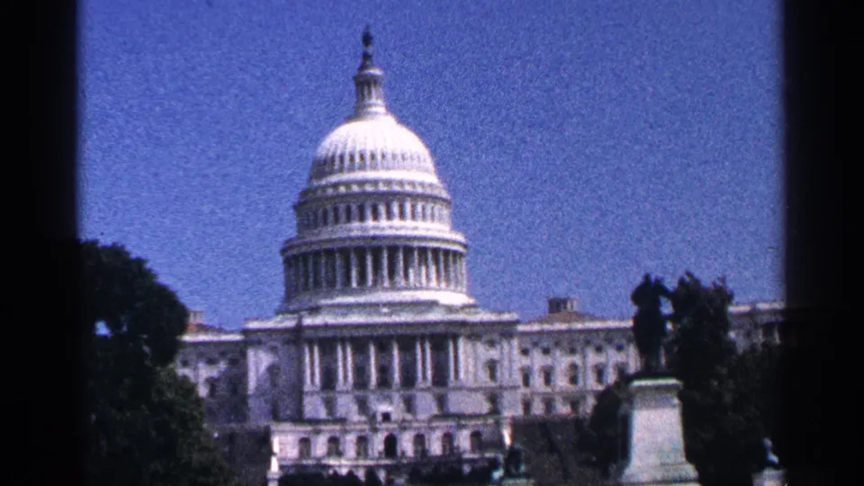1964 8mm view of the U.S. Capitol in Washington DC, framed by trees, a statue silhouette, blue sky, and dark film edges.