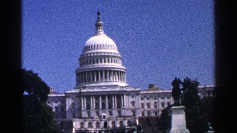 Mid-1960s 8mm frame of the U.S. Capitol in Washington DC, with the dome centered and foreground trees and statue in silhouette.