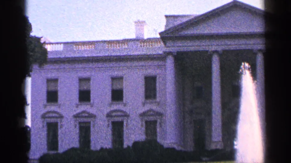 1964 home-movie frame of the White House exterior, with the portico, windows, hedges, and fountain spray softened by 8mm film grain.