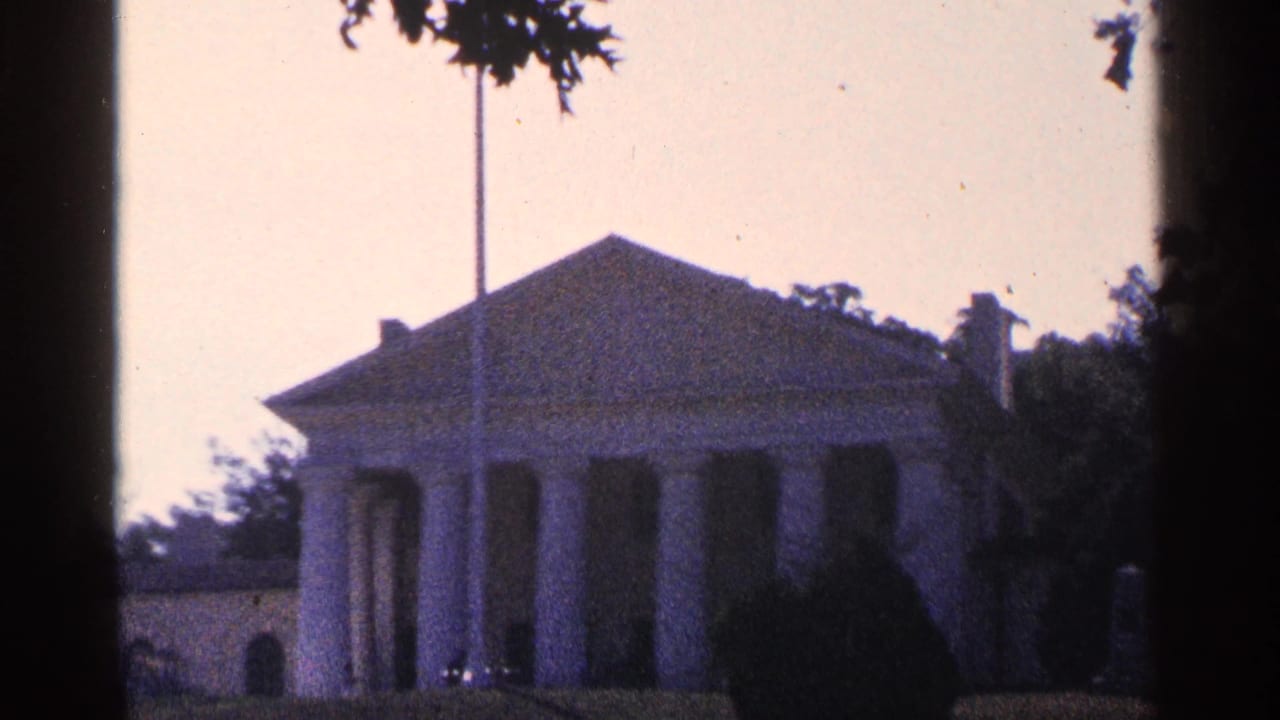 Distant neoclassical house with columns, flagpole, trees, and lawn.