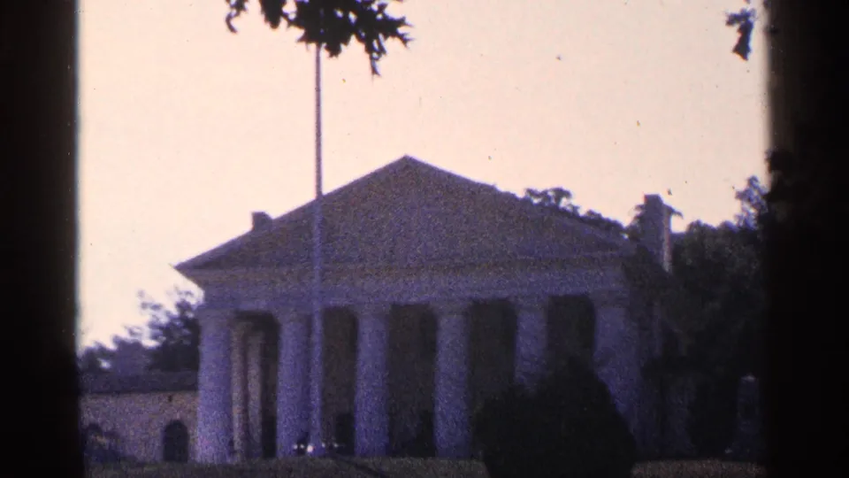 1964 home-movie view of Arlington House from a distance, with columns, a tall flagpole, tree branches, and lawn visible.