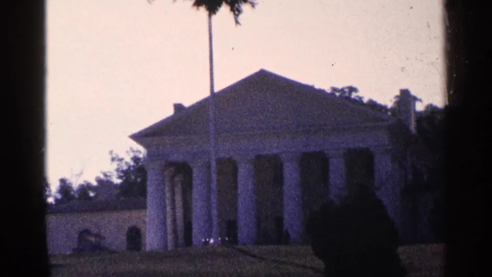 1964 8mm frame showing Arlington House and a flagpole from the lawn, with trees and heavy film-edge shadow in the foreground.