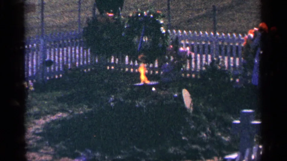 1964 home-movie detail of a memorial wreath with a small flame, set in grass near a white picket fence in a cemetery area.