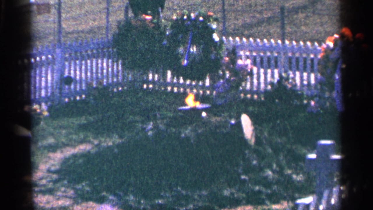 1964 Washington, DC 8mm frame showing an eternal flame, large wreath, white fence, and greenery at a memorial site.