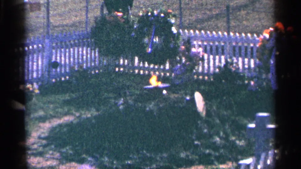 1964 Washington, DC 8mm frame showing an eternal flame, large wreath, white fence, and greenery at a memorial site.