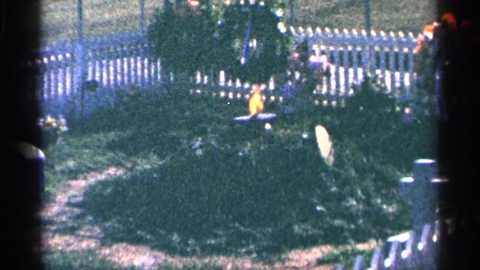 1964 Washington, DC home-movie frame showing a memorial wreath, white fence, shrubs, and a small eternal flame in daylight.