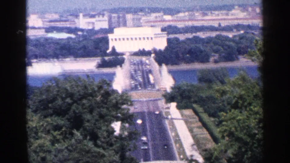1964 Washington DC home-movie frame looking toward the Lincoln Memorial across water and tree-lined roads with traffic below.